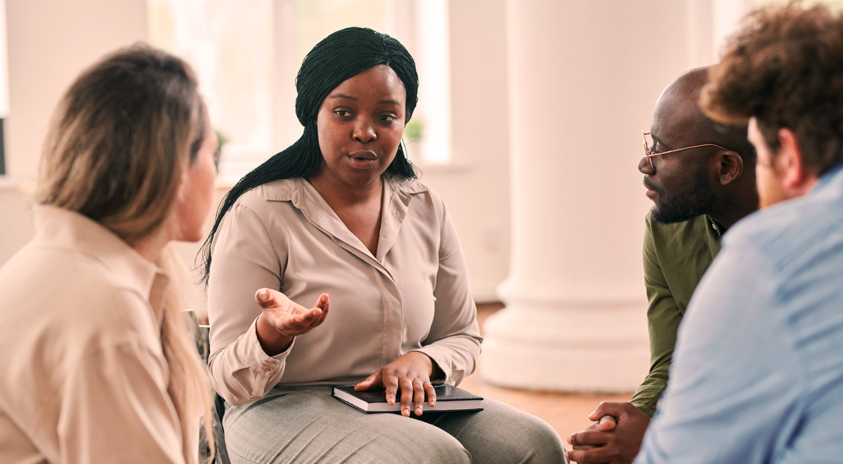 Four people sitting in a circle, two women and two men. One lady talking, the other three are listening.