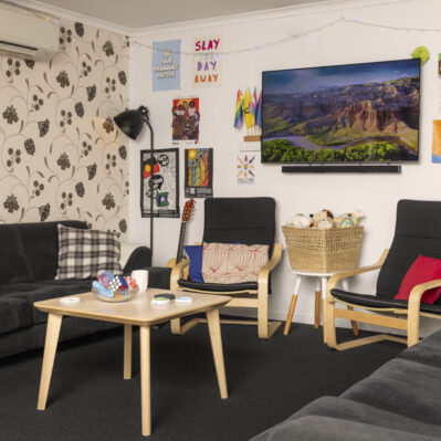 Comfortable couches and armchairs are around a coffee table, on top of which are small sensory toys. Against the wall is a basket of soft toys and a guitar, bright posters and a wall-mounted TV.