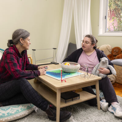 A resident and staff member sit opposite each other on floor pillows. As they talk, the resident holds a soft toy and the staff holds a fidget toy.