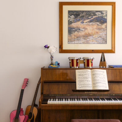 Two guitars lean against a piano. Some flowers, mini conga drums, a metronome and some books sit atop the piano.