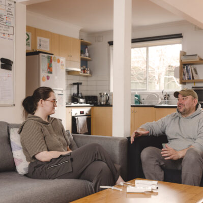 A resident and her peer worker sit on comfortable couches, chatting. The shared kitchen is in the background.