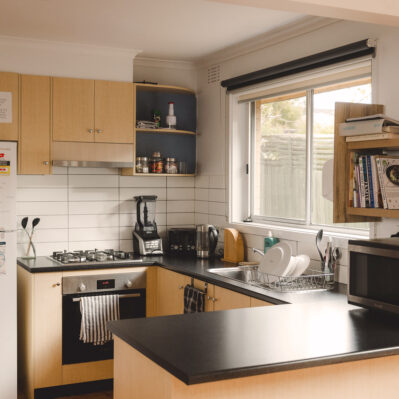 Kitchen contains an oven, stovetop, sink, kettle, blender and microwave. Above the bench are cabinets and shelves containing spices and cookbooks.