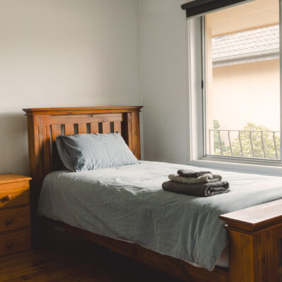 A single bed with a timber bedside table, well-lit by the window beside it. Clean towels are folded on top of the neatly made sheets.