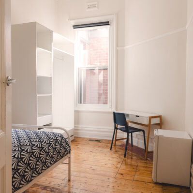 A view of the room from the doorway. At the foot of the bed is an empty wardrobe and shelves. Against the opposite wall is a small desk, office chair and mini fridge.