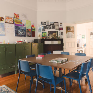 Eight chairs surround a table, on top of which is a puzzle in progress and containers of jewellery beads. Against the walls are cabinets and artworks by the residents.