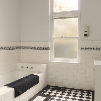 A bath sits against the wall adjacent to a frosted glass window and opposite the sink. Chequered black and white tiles cover the floor.