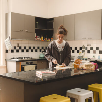 A resident making herself a sandwich. Kitchen includes an oven and stove top, standard appliances, cabinets and shelves holding condiments and other goods.