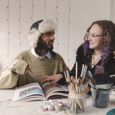 Two residents sitting at a table, flipping through a magazine and smiling. Paint brushes, washi tape and other art materials are set out in front of them.