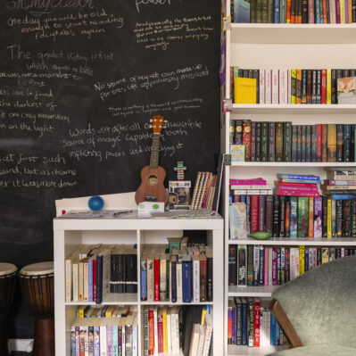 Well-stocked bookshelves stand in front a large blackboard showing reflections from the residents written in different coloured chalk. By the shelves are two djembes and a ukulele.