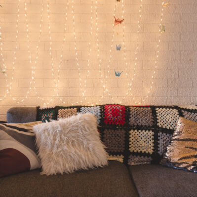 A cozy couch with fairly lights and paper cranes decorating the wall behind it. Cushions of different textures and a crocheted throw decorate the couch.