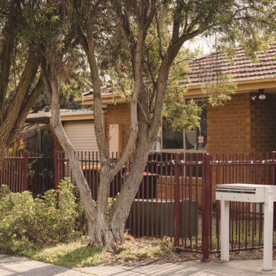 A brick unit with a fenced yard. In front of the fence are mailboxes, native trees and flowers.
