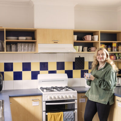 Resident Bree stands in her kitchen, smiling with a cup of tea. Cookbooks, glasses, mugs, tea and coffee are stacked neatly on the shelves behind her. The kitchen includes an oven, fridge, sink and standard appliances.