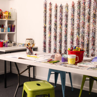 Paint brushes, pencils, markers and other art materials are spread across a long table. There is a small activity table and storage caddy containing paints and more materials in the corner. Decorating the wall are hundreds of multicoloured paper cranes strung together.