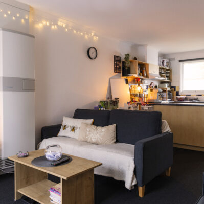 Living room features sofas arranged around a small coffee table, storage cubes containing books and a TV. The living space backs onto the kitchen area.