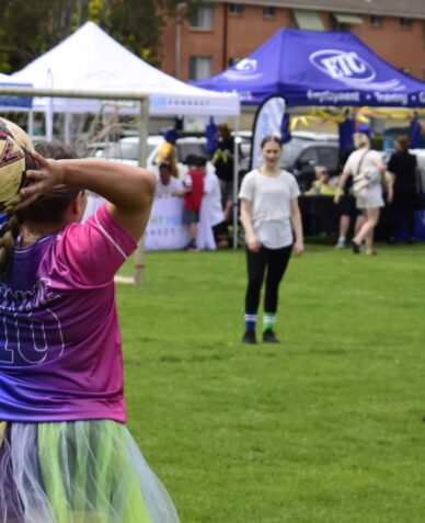 A person wearing a tutu prepares to throw-in a soccer ball to other players, information stall in the background.