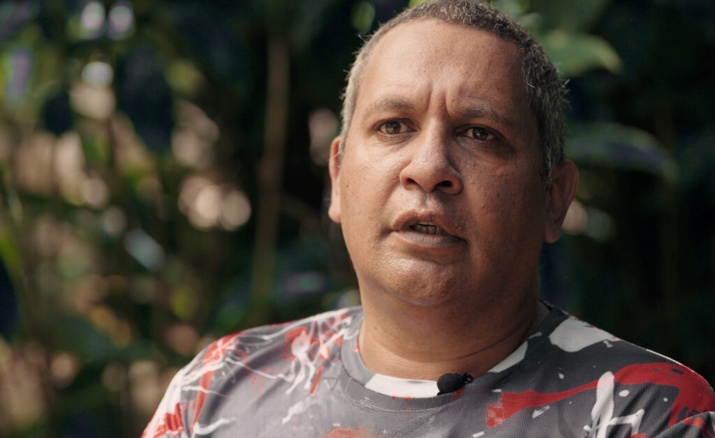 Warren is looking at someone standing beside the camera and sitting outside. Plants can be seen in the background