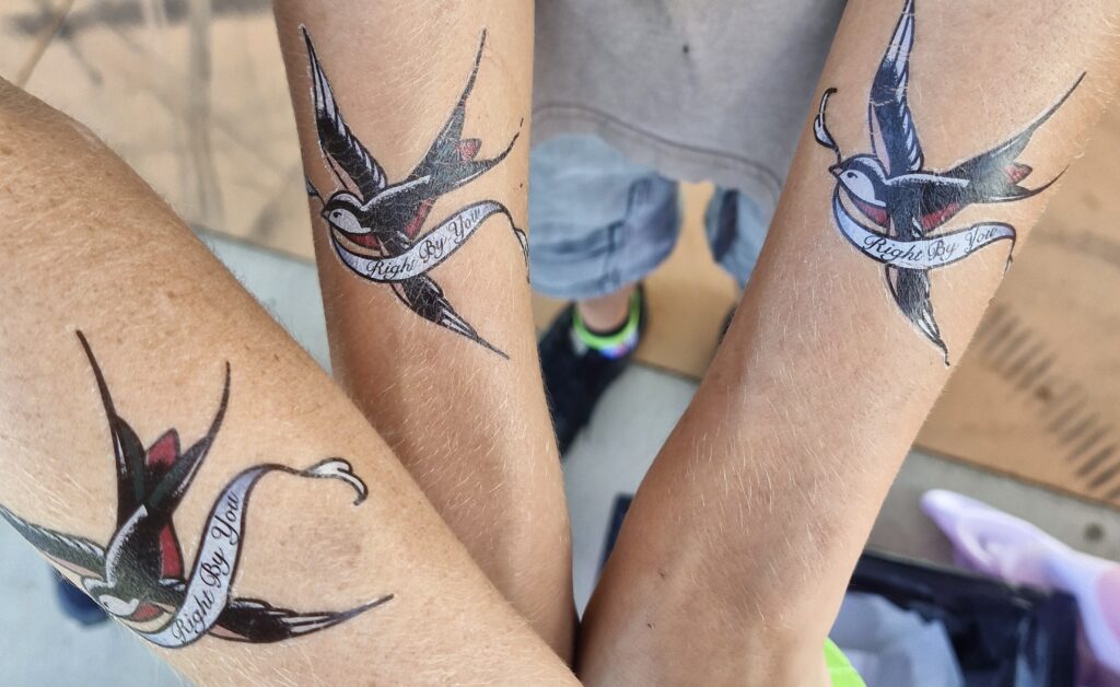 Two people hold out their forearms, showing off a temporary traditional-style tattoo of a black swallow carrying a banner saying 'RightByYou' in cursive.