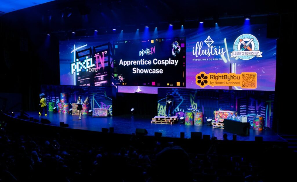 The main stage of Pixel Expo, set up with blue lighting and a large screen behind it. In the centre of the screen, it says 'Apprentice Cosplay Showcase' and to the left and right are the Pixel Expo logos and various sponsors, including RightByYou.